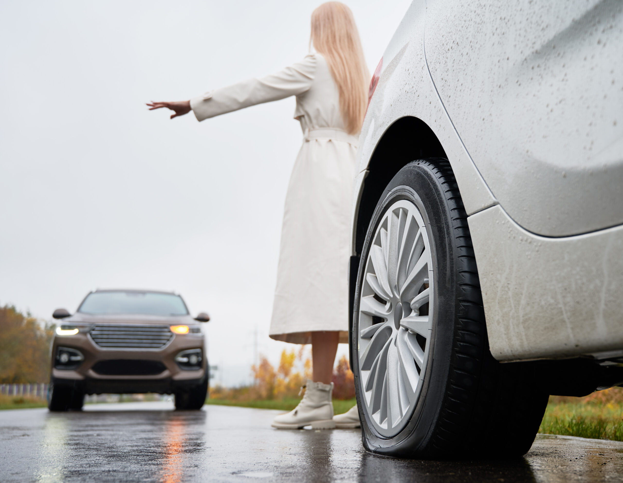 Back view of girl standing behind white vehicle with rear flat tire parked on edge of roadway and trying to stop oncoming SUV. Female driver of auto with damage tire giving stop sign to oncoming car.