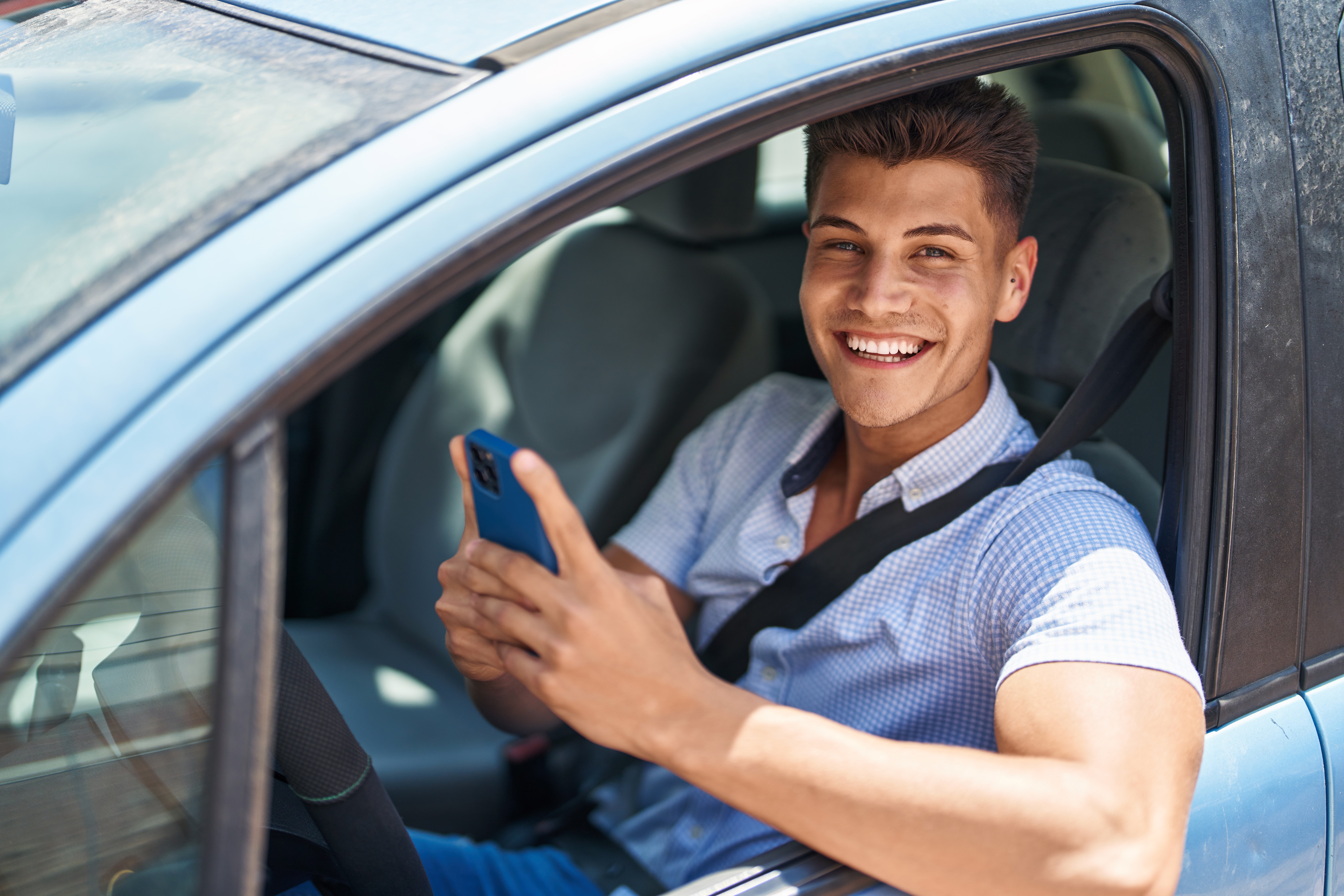 Young hispanic man using smartphone sitting on car after getting fuel delivered