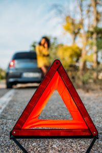 A red warning triangle is placed on the road, with a blurred car and a person beside it in the background, indicating a roadside emergency or breakdown that may require roadside assistance.